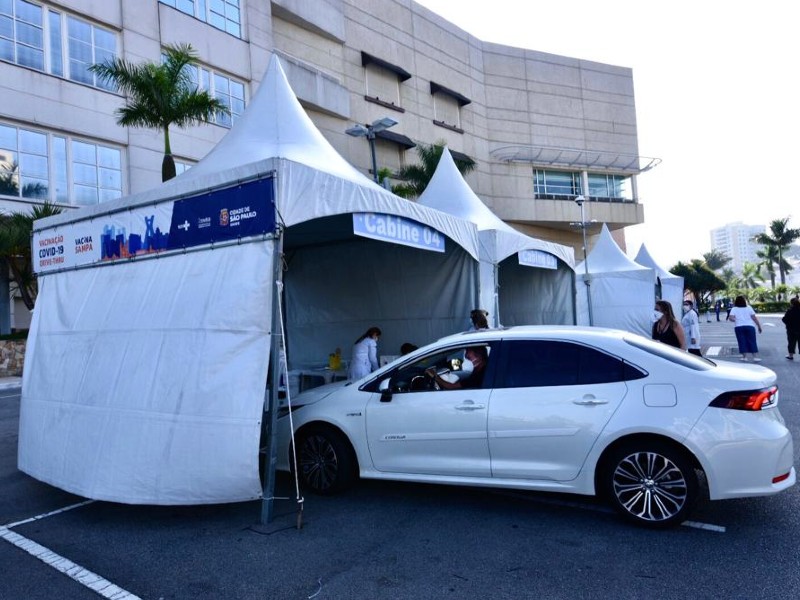 Foto do drive-thru do Shopping Anália Franco. Em primeiro plano estão quatro tendas onde acontece a vacinação. Na primeira tenda há um carro branco estacionado com uma pessoa sendo vacinada. O prédio do shopping aparece ao fundo